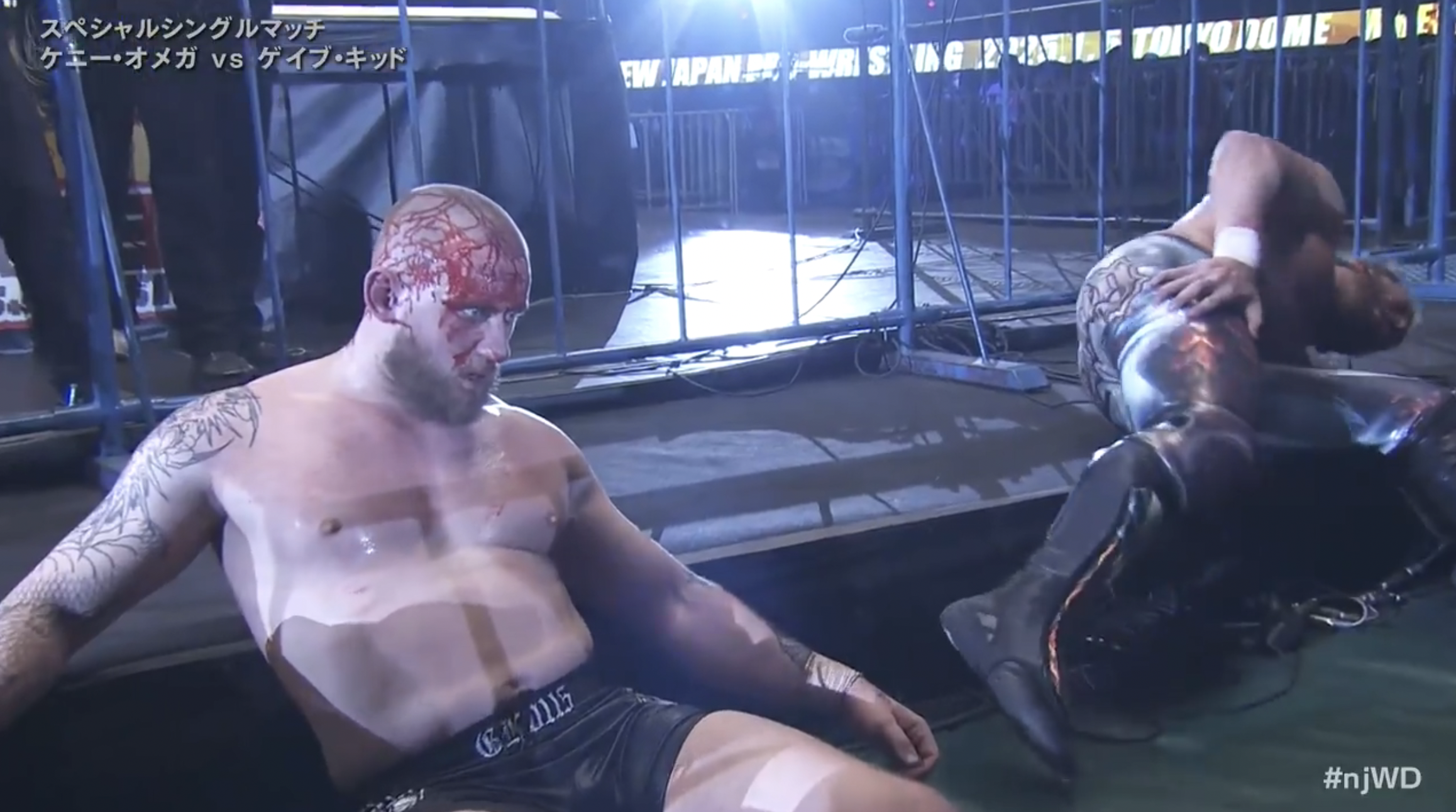 Gabe Kidd stares ahead, covered in blood, as Kenny Omega holds his hip and writhes in pain.